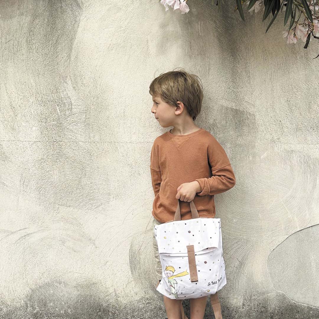 Kind staat naast een muur en houdt de Petit Jour schoolrugzak met De Kleine Prins in de hand.