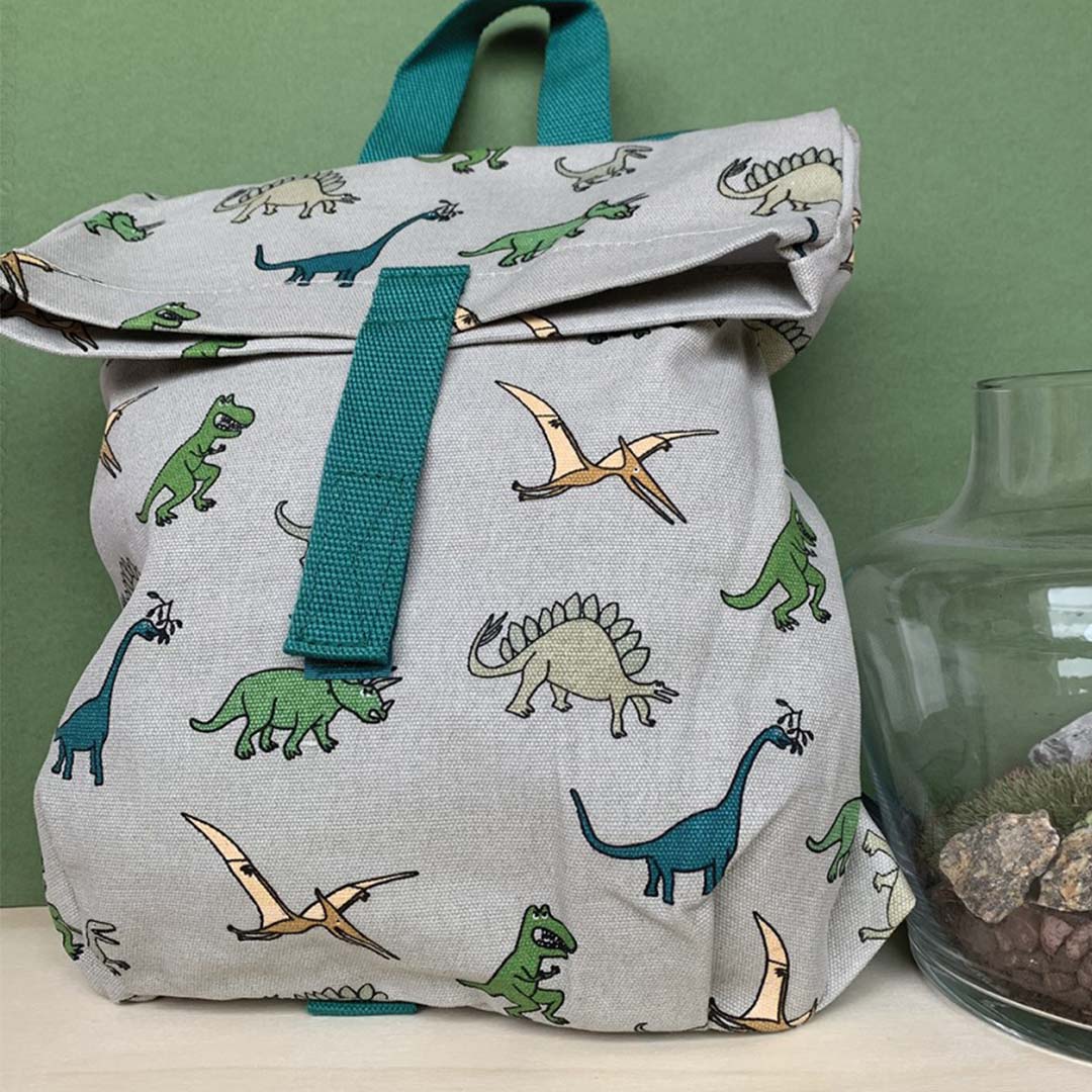 petit jour schoolrugzak met dinosauruspatroon, schuin van voren gefotografeerd met een glazen object op de achtergrond.