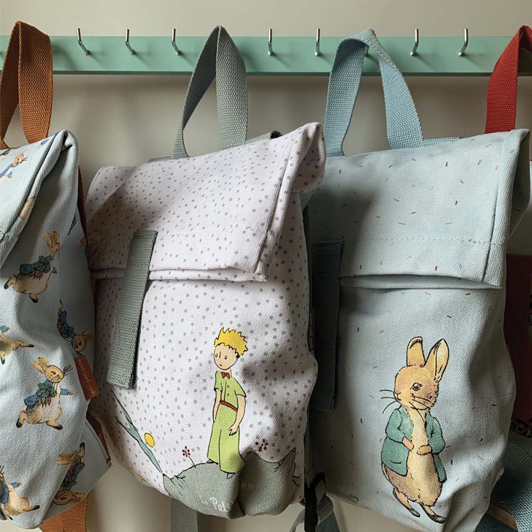 Drie Petit Jour kinderrugzakken, waaronder twee met Pieter Konijn en één met Le Petit Prince, hangend aan een kapstok.