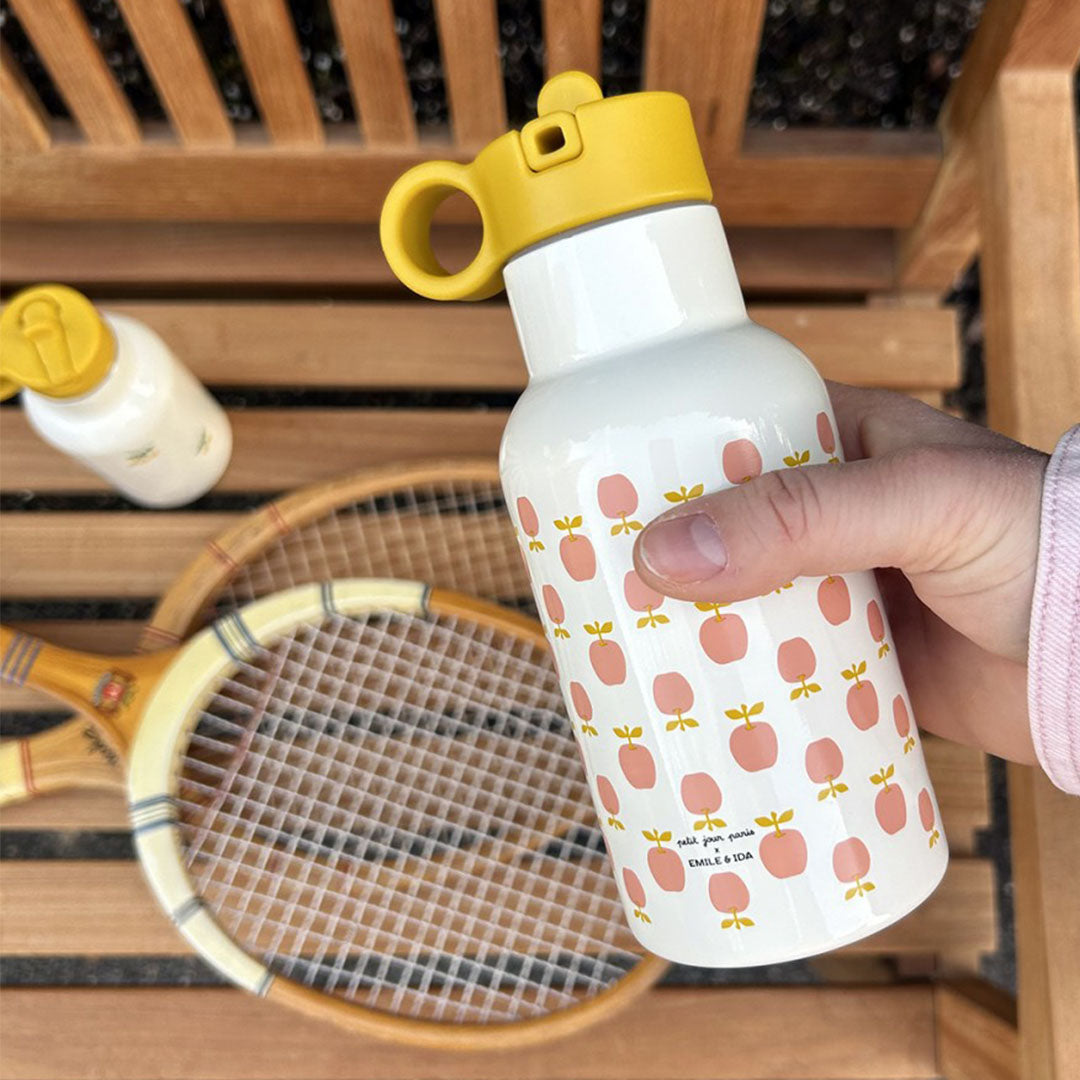 Een hand houdt een witte rvs drinkfles met appelpatroon vast, op een houten bank met een tennisracket.