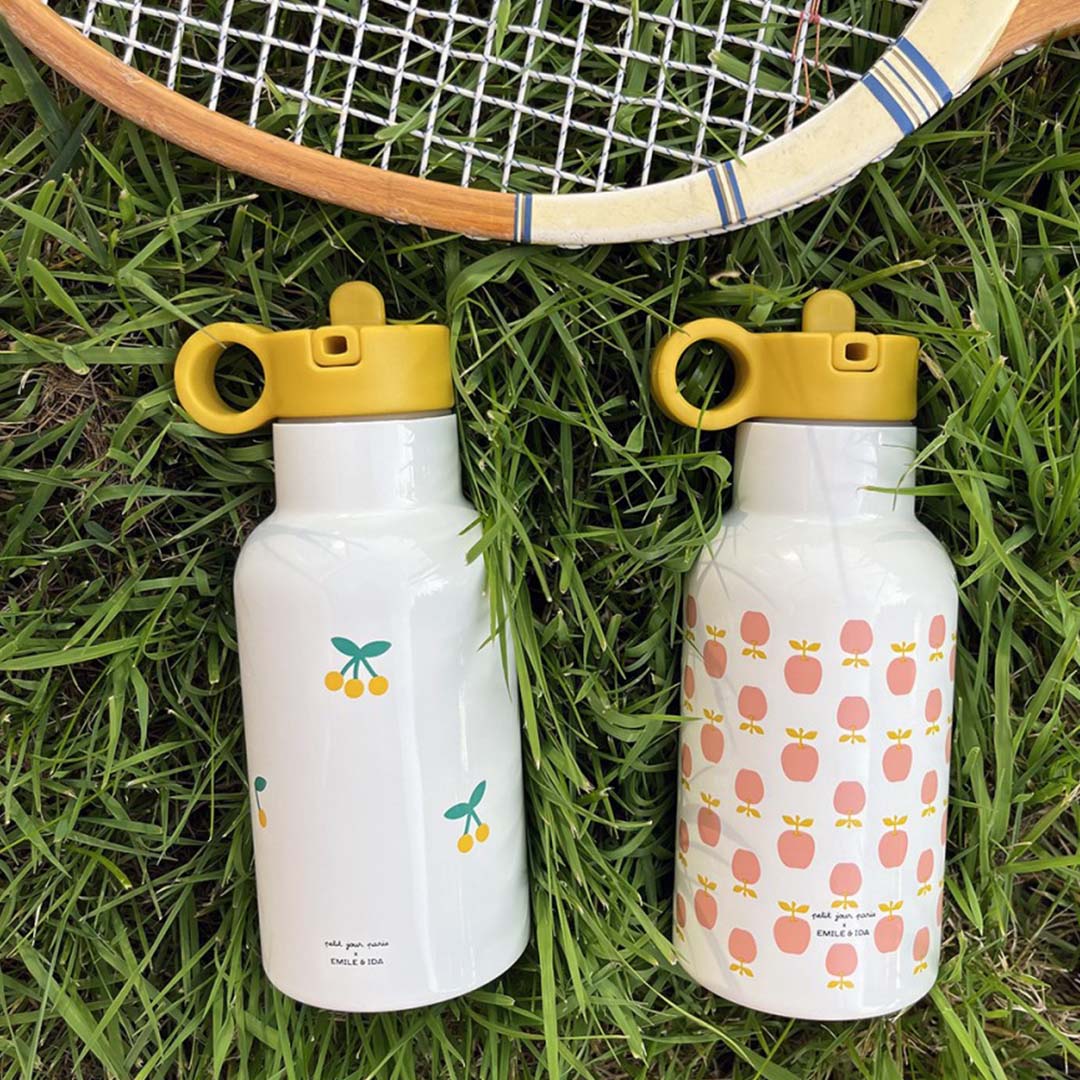 Twee witte rvs drinkflessen, één met appelpatroon en één met kersenpatroon, liggen op gras naast een tennisracket.