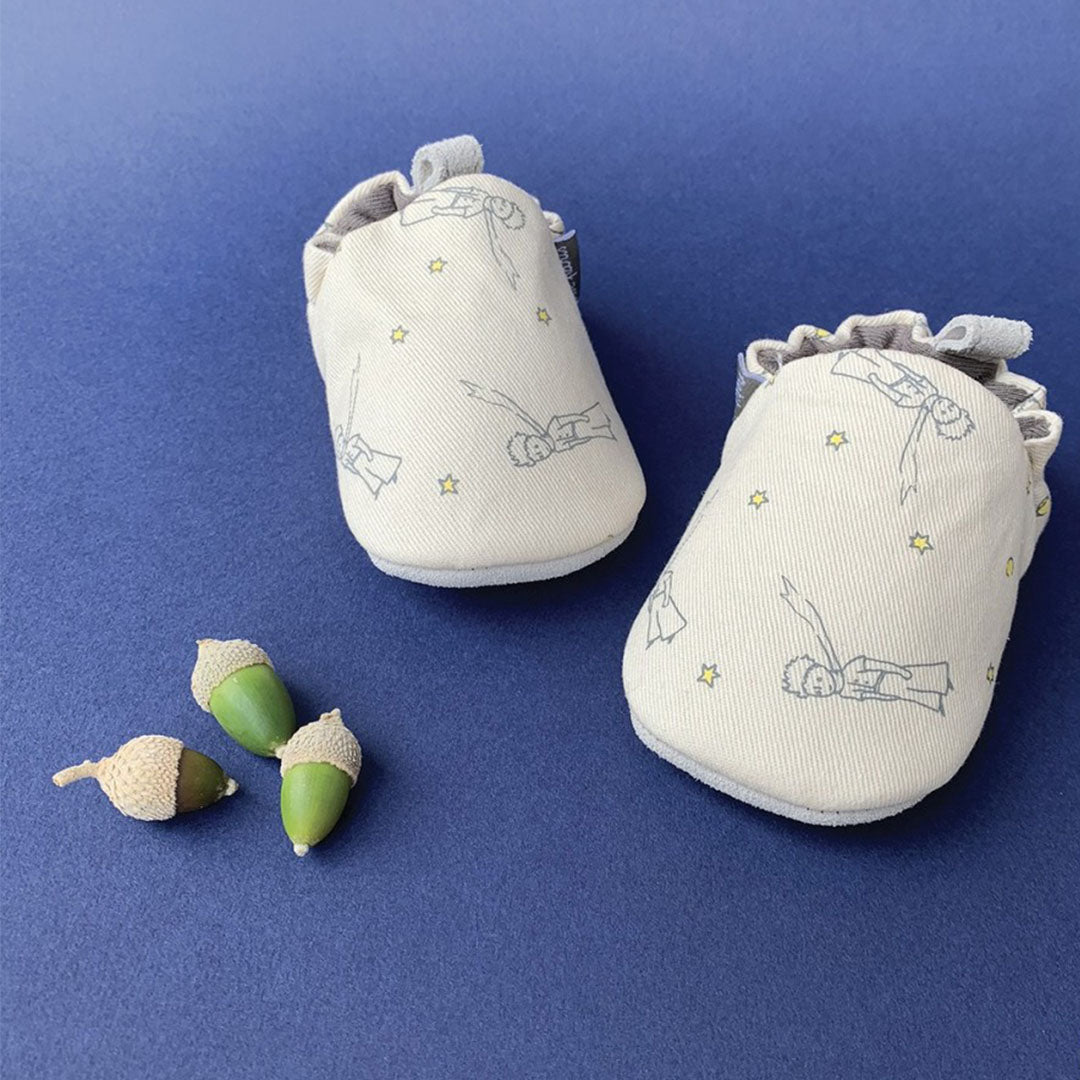 Twee petit jour baby schoentjes met zachte zool, beige met een patroon van De Kleine Prins en sterren, op een blauwe ondergrond met decoratieve eikels.
