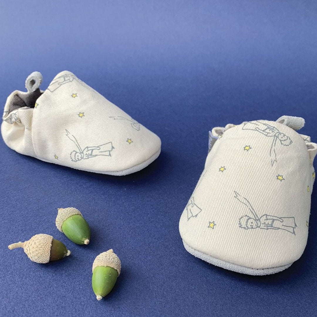 Twee petit jour baby schoentjes met zachte zool, beige met een patroon van De Kleine Prins en sterren, op een blauwe ondergrond met decoratieve eikels.