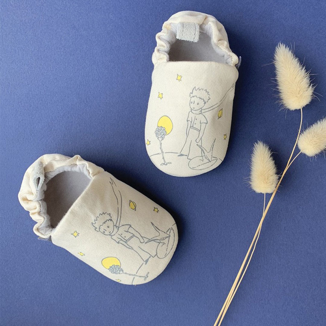 Twee beige petit jour baby schoentjes met zachte zool, bedrukt met Le Petit Prince en sterren, op een blauwe ondergrond.