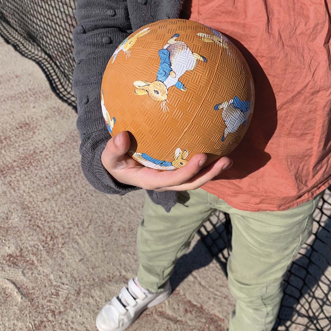 Een kind houdt de oranje Petit Jour speelbal met Pieter Konijn illustraties vast.