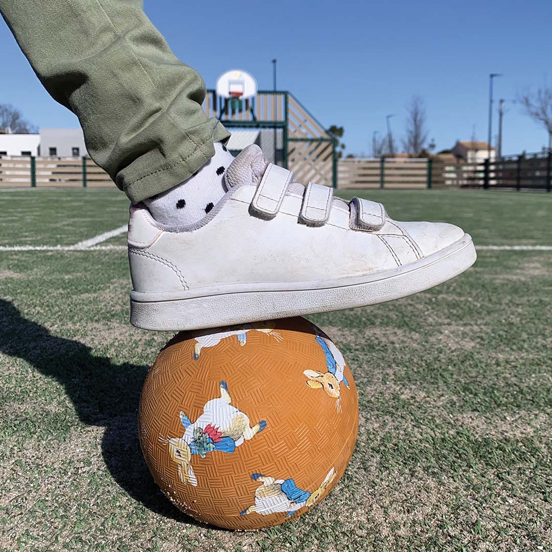 Een kindervoet in een witte sneaker staat op de oranje Petit Jour speelbal met Pieter Konijn op een sportveld.