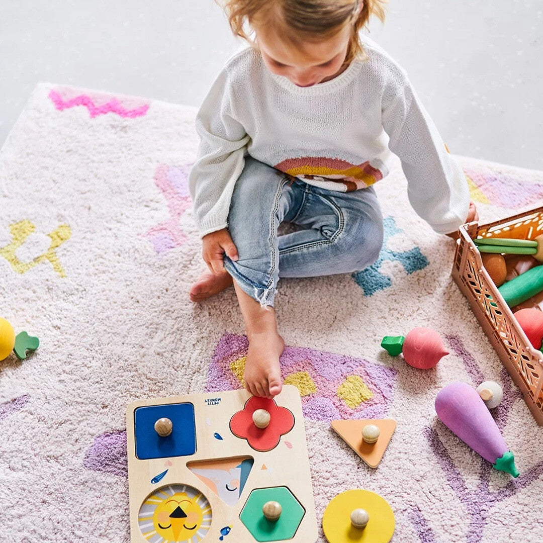 Een kind speelt op een vloerkleed met de Petit Monkey houten vormenpuzzel 'One day at the Zoo' en houten speelgoedgroenten.