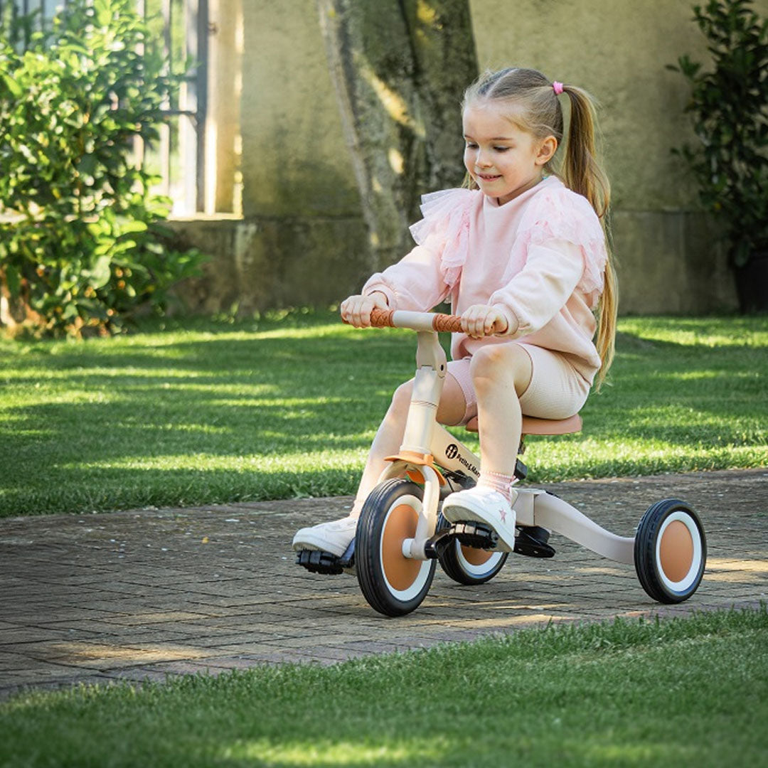 Een lachend meisje rijdt op de Petite&Mars 5-in-1 kinderfiets in driewielerstand op een pad in de tuin.