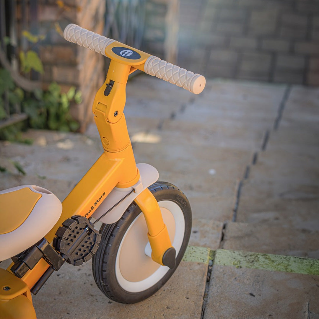 Detail van het stuur en voorwiel van de gele Petite&Mars kinderfiets op een stenen ondergrond.