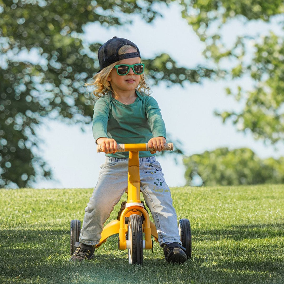 Een kind met zonnebril en pet rijdt op de gele Petite&Mars kinderfiets over gras.