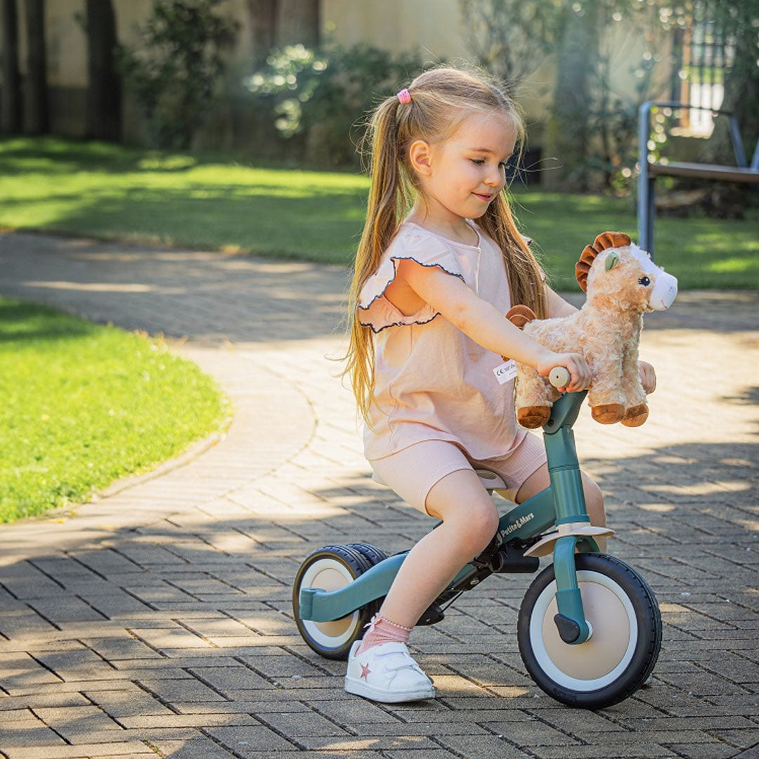 Een meisje zit op de groene Petite&Mars 5-in-1 kinderfiets in loopfietsstand met een speelgoedpaard.