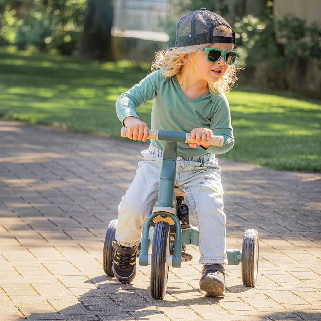 Een kind rijdt op de groene Petite&Mars 5-in-1 kinderfiets in driewielerstand met pedalen op een pad.