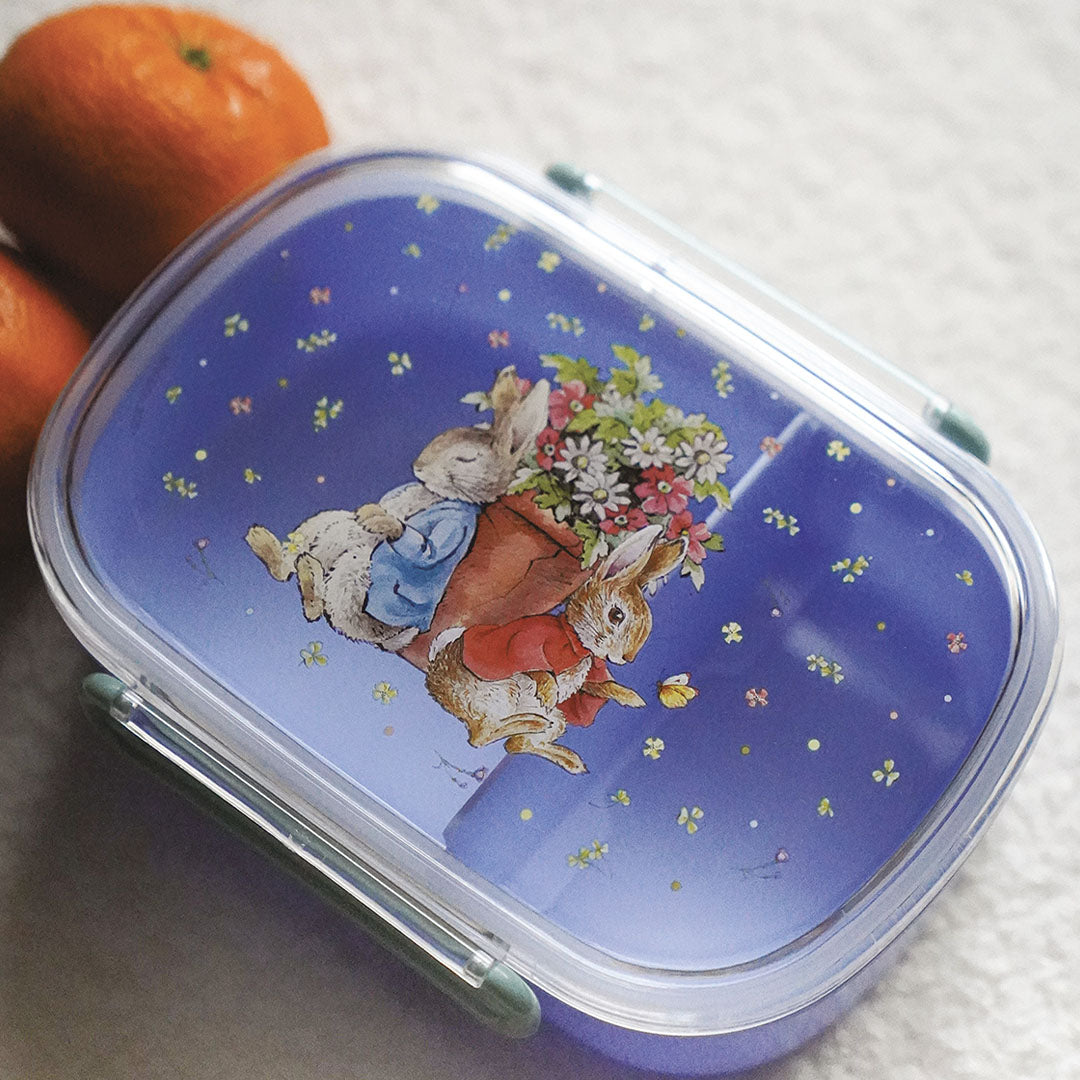 Petit Jour kinderbroodtrommel met Pieter Konijn illustratie op de deksel, gefotografeerd met fruit op de achtergrond.