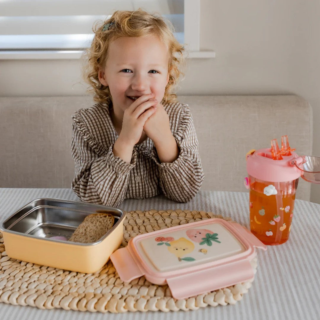 Lachend kind aan tafel met een open petit monkey bento broodtrommel en een drinkfles.