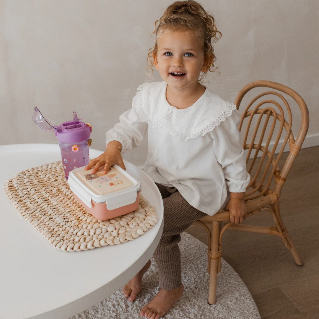 Een lachend kind zit aan tafel met de petit monkey bento broodtrommel en een paarse drinkfles.