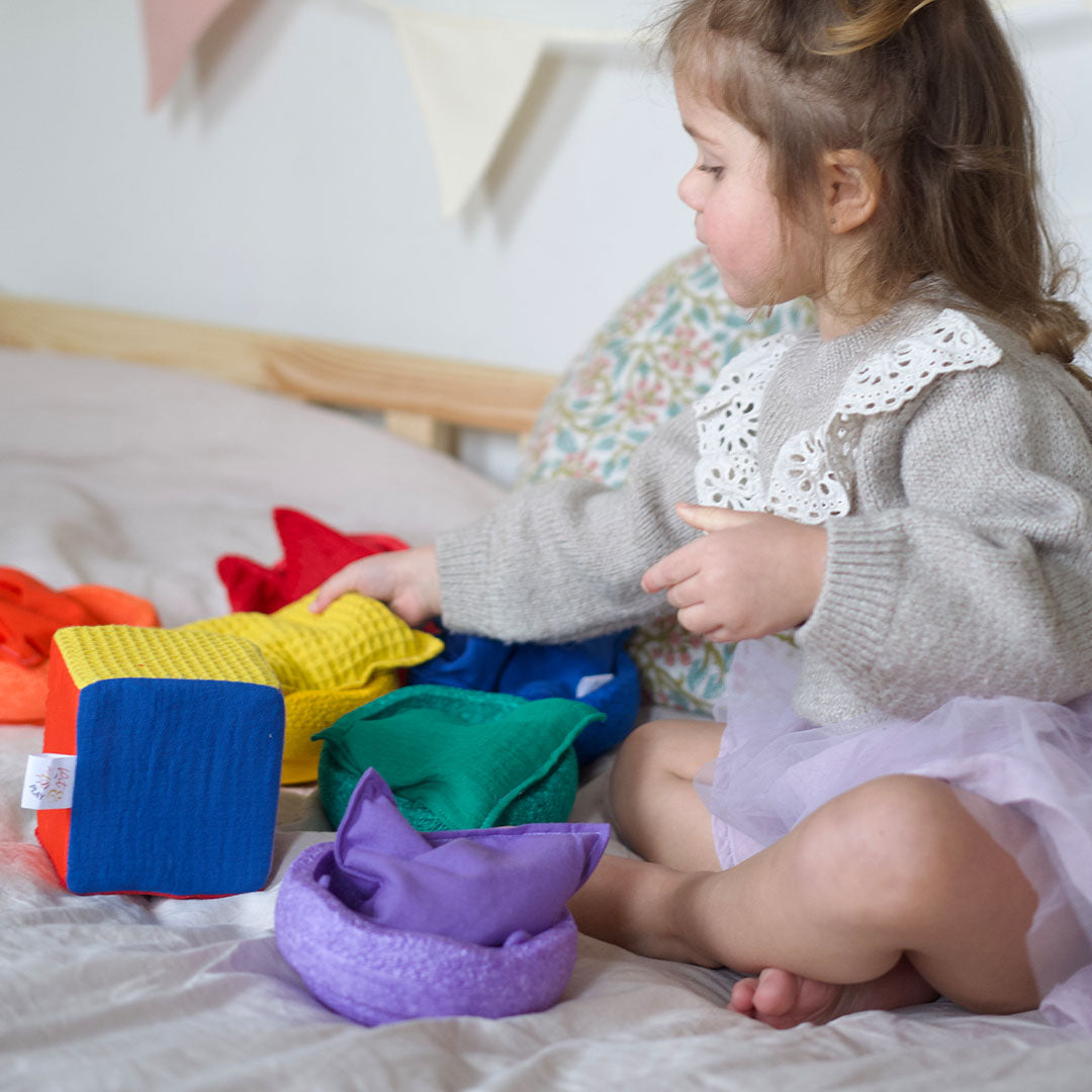 Een kind speelt op bed met een veelkleurige stoffen dobbelsteen en bijpassend sensorisch speelgoed.