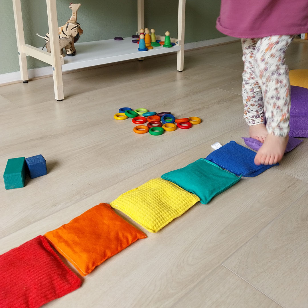 Een kind stapt met blote voeten over een rij regenboogkleurige pittenzakken op een houten vloer.
