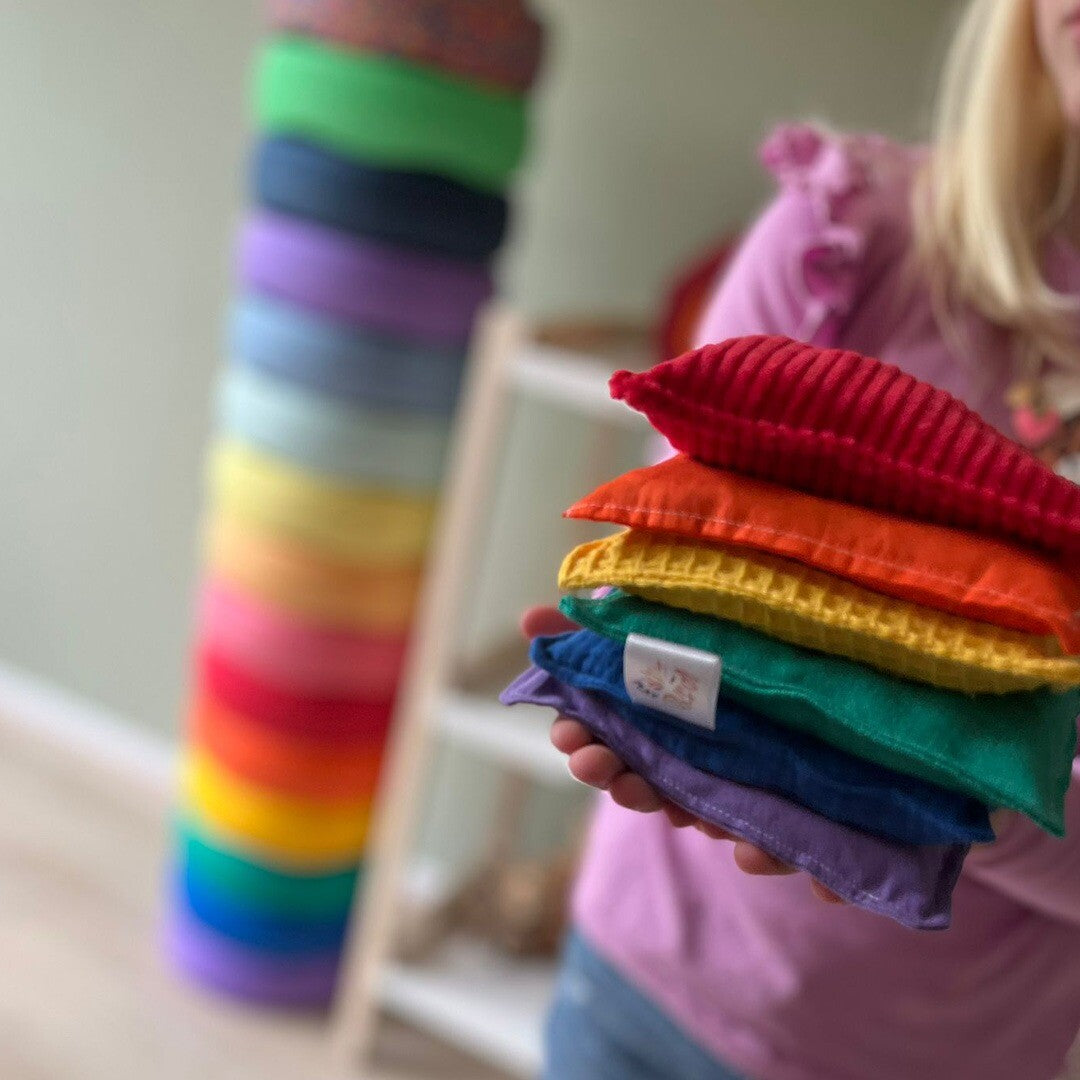Een kind houdt een stapel van zes regenboogkleurige pittenzakken met verschillende texturen vast.