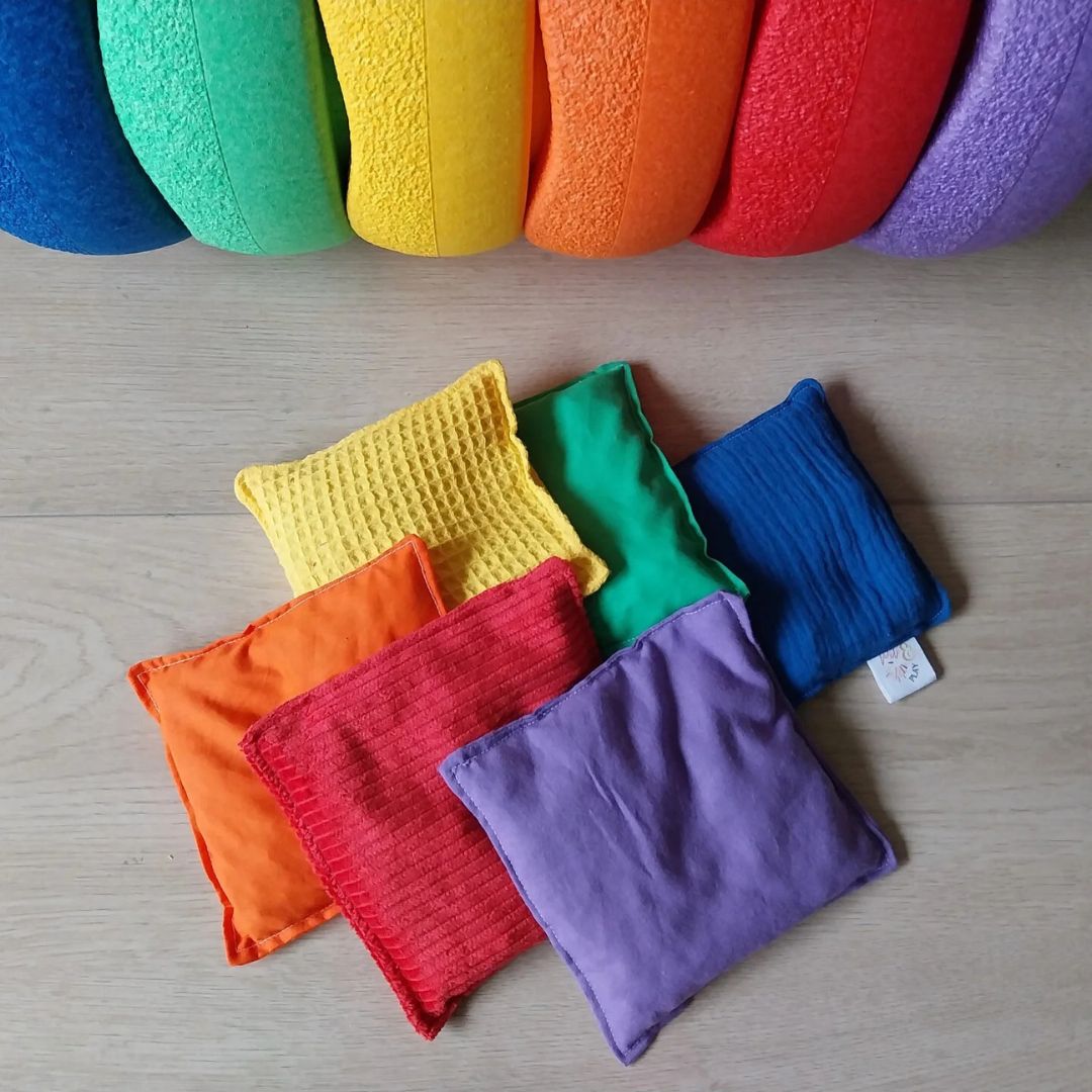 Zes pit and play sensorische pittenzakken in regenboogkleuren en verschillende texturen liggen verspreid op een houten vloer.