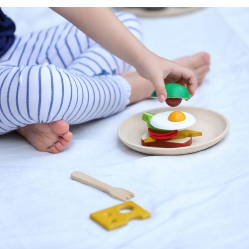 Een kind speelt met de Plan Toys houten ontbijtset op een witte deken.