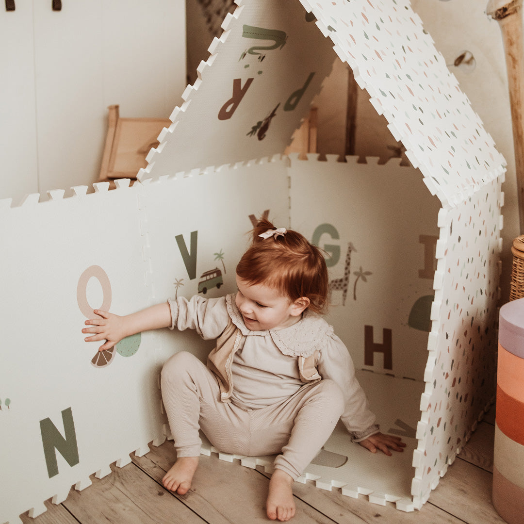 Een peuter zit in een speelhuisje gemaakt van play&go foam speelmat tegels met alfabetletters.