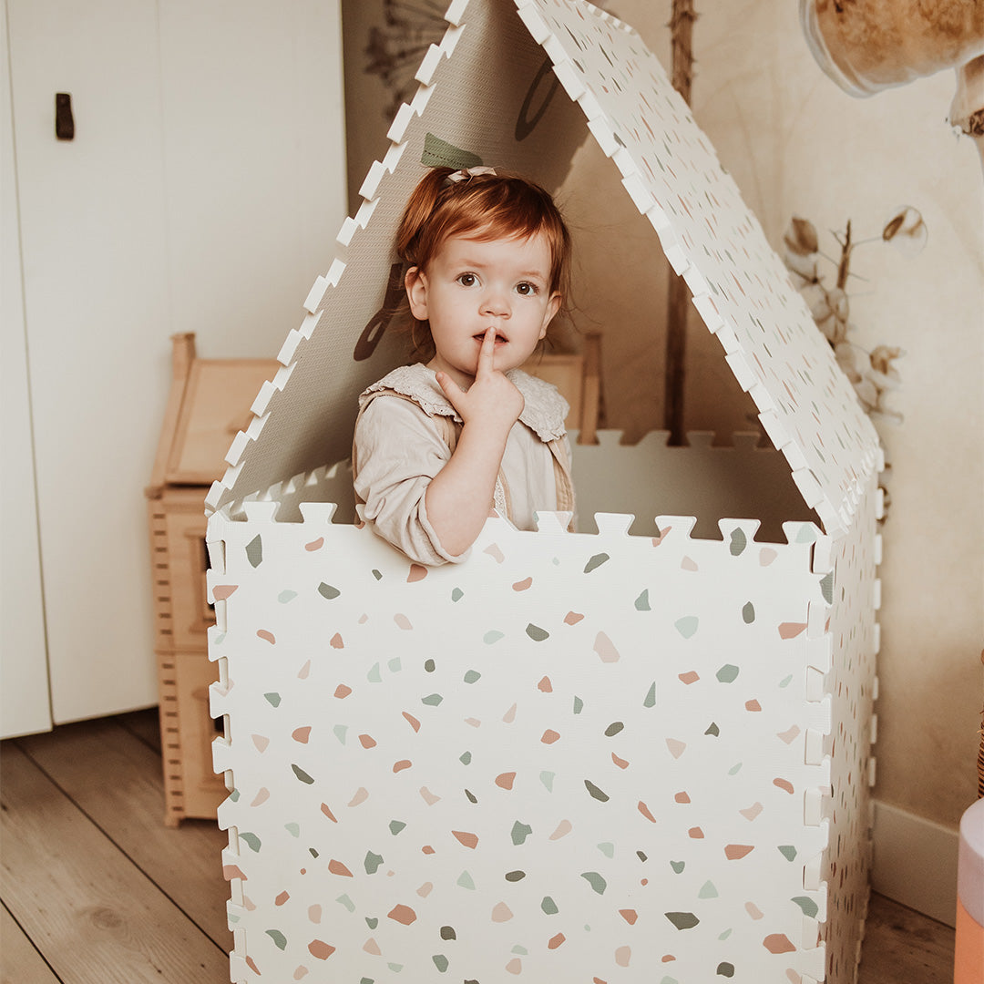 Een peuter kijkt uit een speelhuisje gemaakt van play&go foam speelmat tegels met een willekeurig patroon.