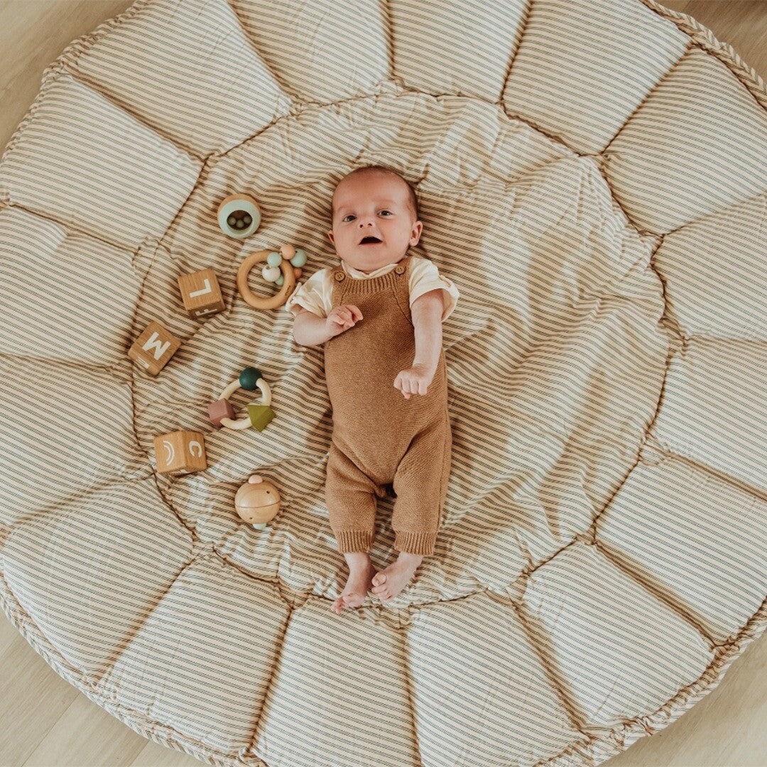 Baby ligt op het uitgevouwen play&go 3-in-1 speelkleed, beige met blauwe strepen, omringd door houten babyspeelgoed.