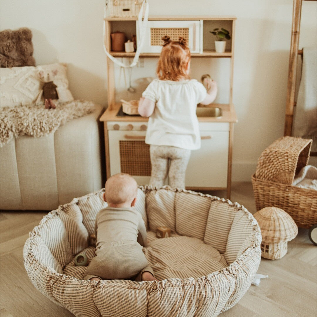 Baby zit in het play&go 3-in-1 speelkleed, kijkend naar een kind dat speelt met een houten speelkeuken.