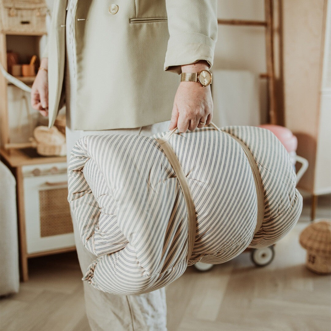 Een persoon draagt het opgerolde en opgevouwen play&go 3-in-1 speelkleed, beige met blauwe strepen, aan de draagbanden.