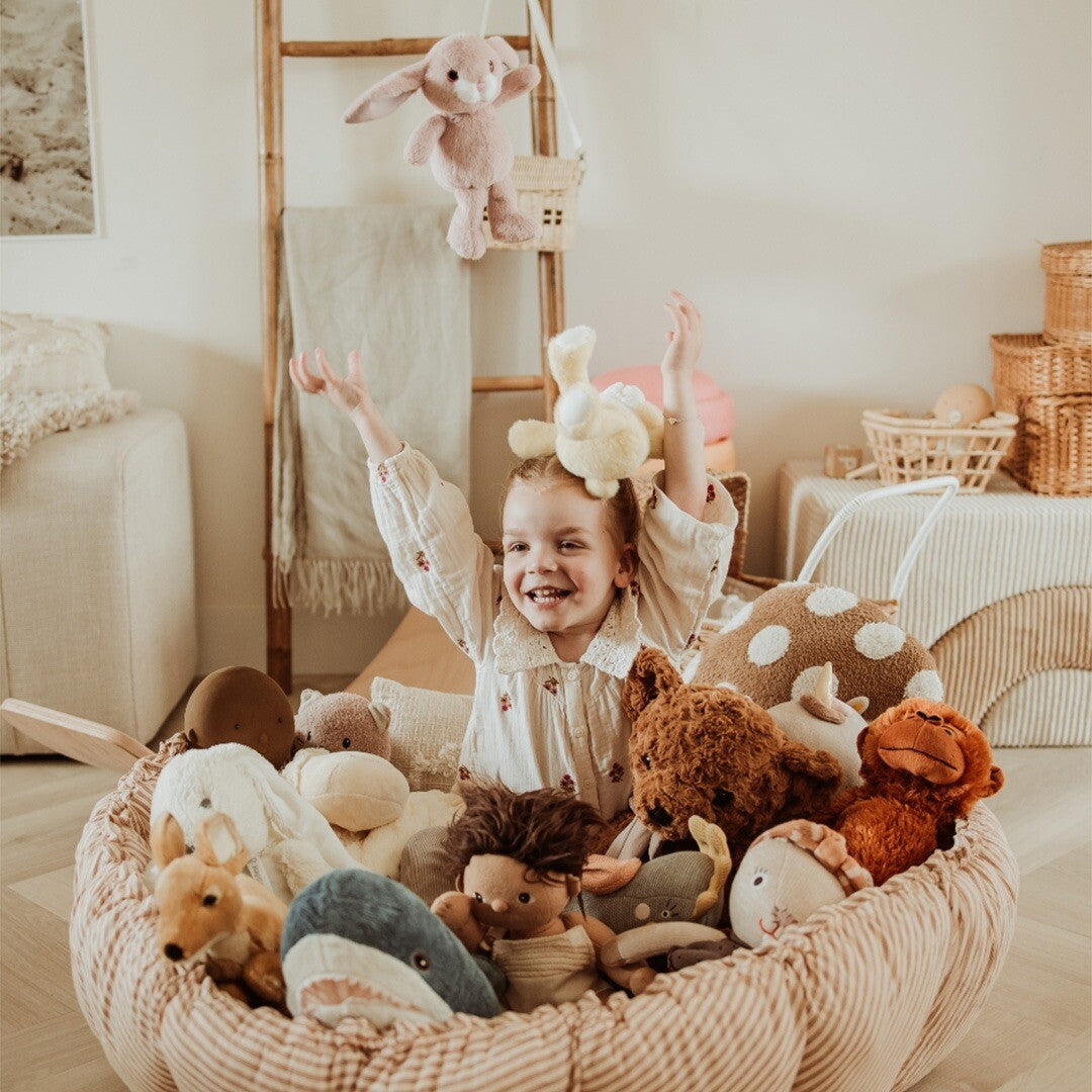 Lachend kind zit in het play&go speelkleed dat is opgevouwen tot een opbergmand vol knuffels.