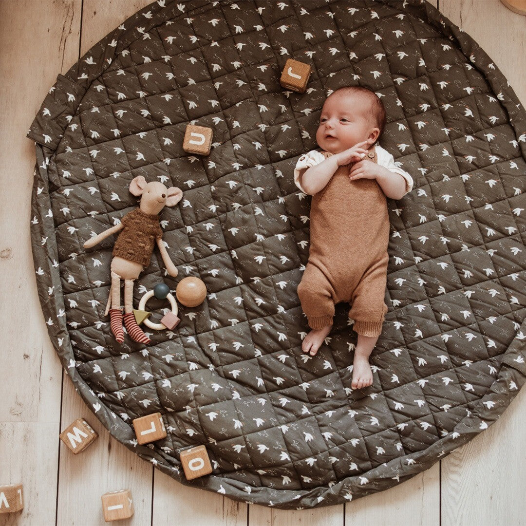 Baby ligt op een play&go groen speelkleed met vogelpatroon, omringd door speelgoed.