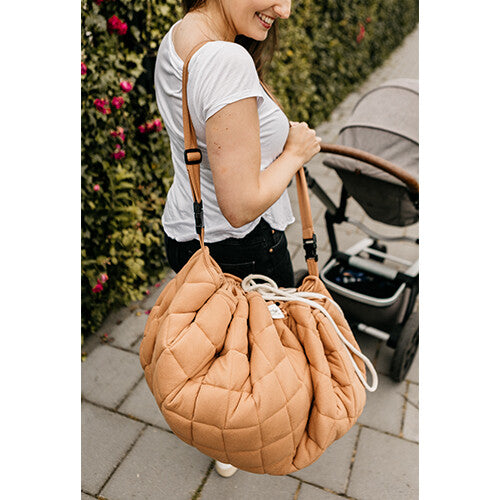 Een persoon draagt het Play&go tawny bruine gewatteerde babyspeelkleed als schoudertas naast een kinderwagen.