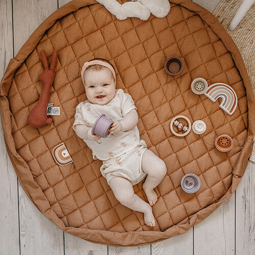 Een lachende baby ligt op het Play&go tawny bruine gewatteerde babyspeelkleed tussen diverse speeltjes.