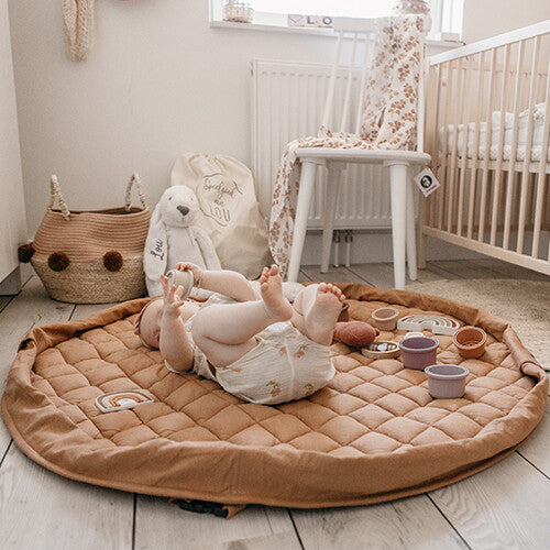Een baby ligt op de rug op het Play&go tawny bruine gewatteerde babyspeelkleed in een babykamer.