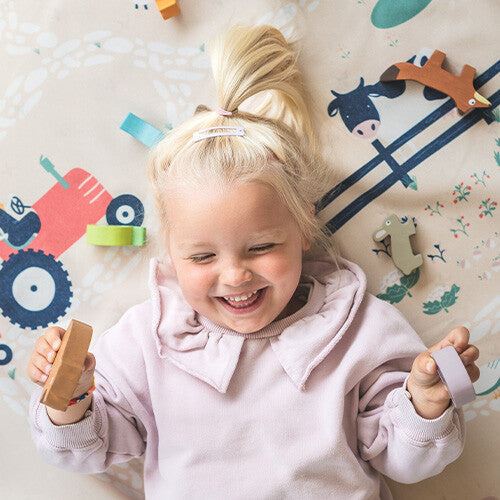 Een lachend kind ligt op het play&go speelkleed met boerderijthema en speelt met houten speelgoed.