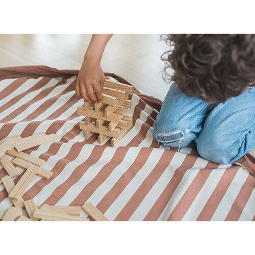 Een kind bouwt met houten blokken op het open, bruin-wit gestreepte speelkleed.