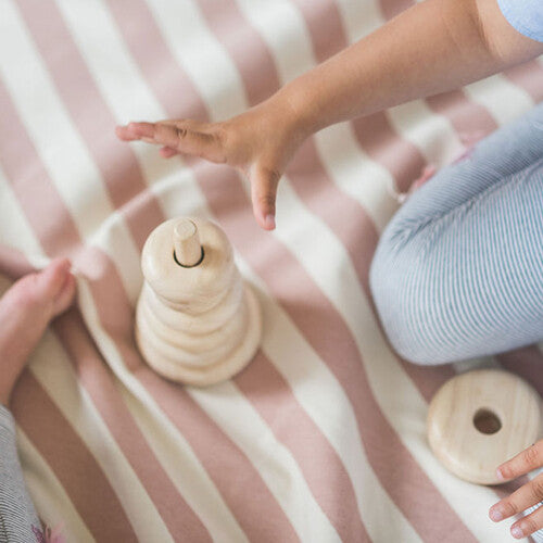 Detail van kinderhanden die spelen met een houten stapeltoren op een play&go speelkleed met roze en witte strepen.