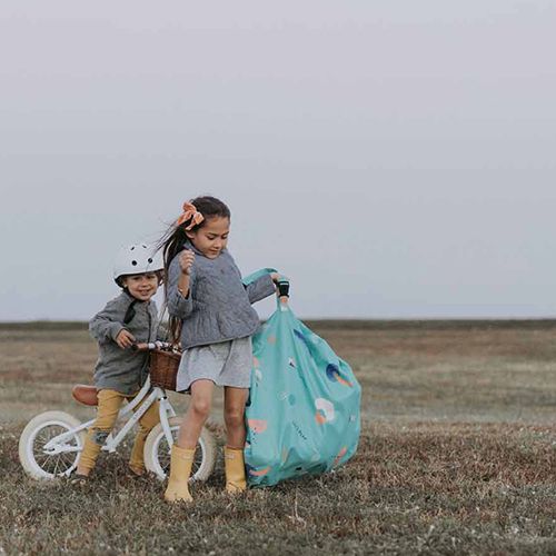 Twee kinderen staan in een grasveld, waarvan één de play&go waterdichte speelkleed en opbergzak draagt en de ander naast een loopfiets staat.
