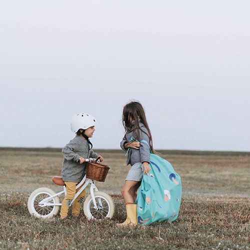 Twee kinderen staan in een grasveld, waarvan één de play&go waterdichte speelkleed en opbergzak draagt en de ander naast een loopfiets staat.