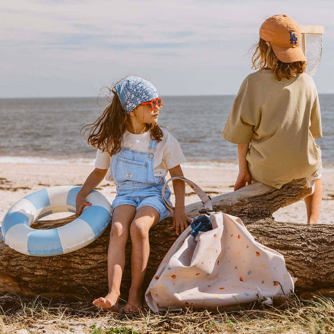 Twee kinderen zitten op een boomstam op het strand met de play&go waterdichte speelkleed en opbergzak ernaast.
