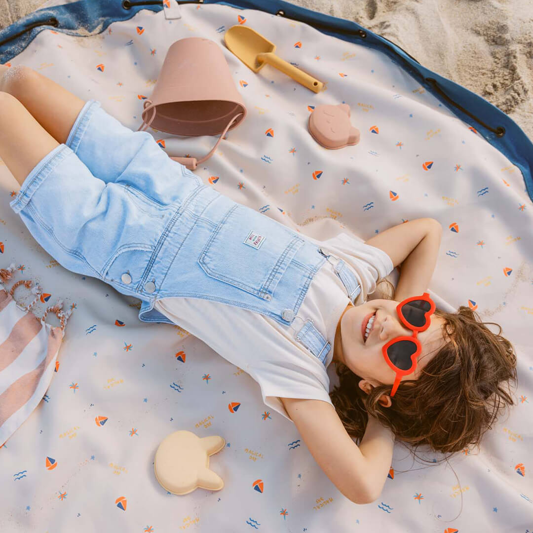 Een kind ligt lachend op het opengevouwen play&go waterdichte speelkleed met Magical Day patroon op het strand.