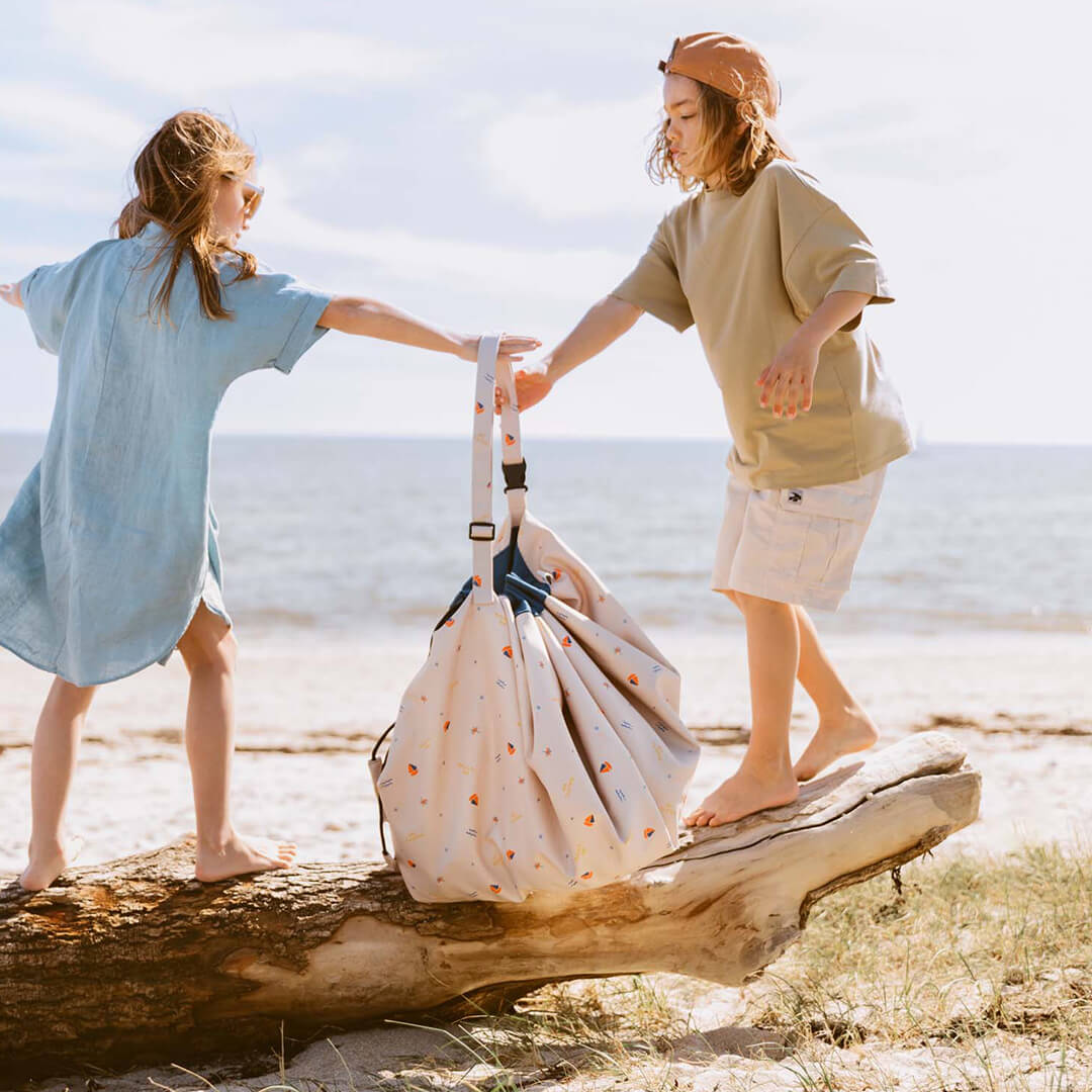 Twee kinderen dragen de play&go waterdichte speelkleed en opbergzak als tas op het strand.