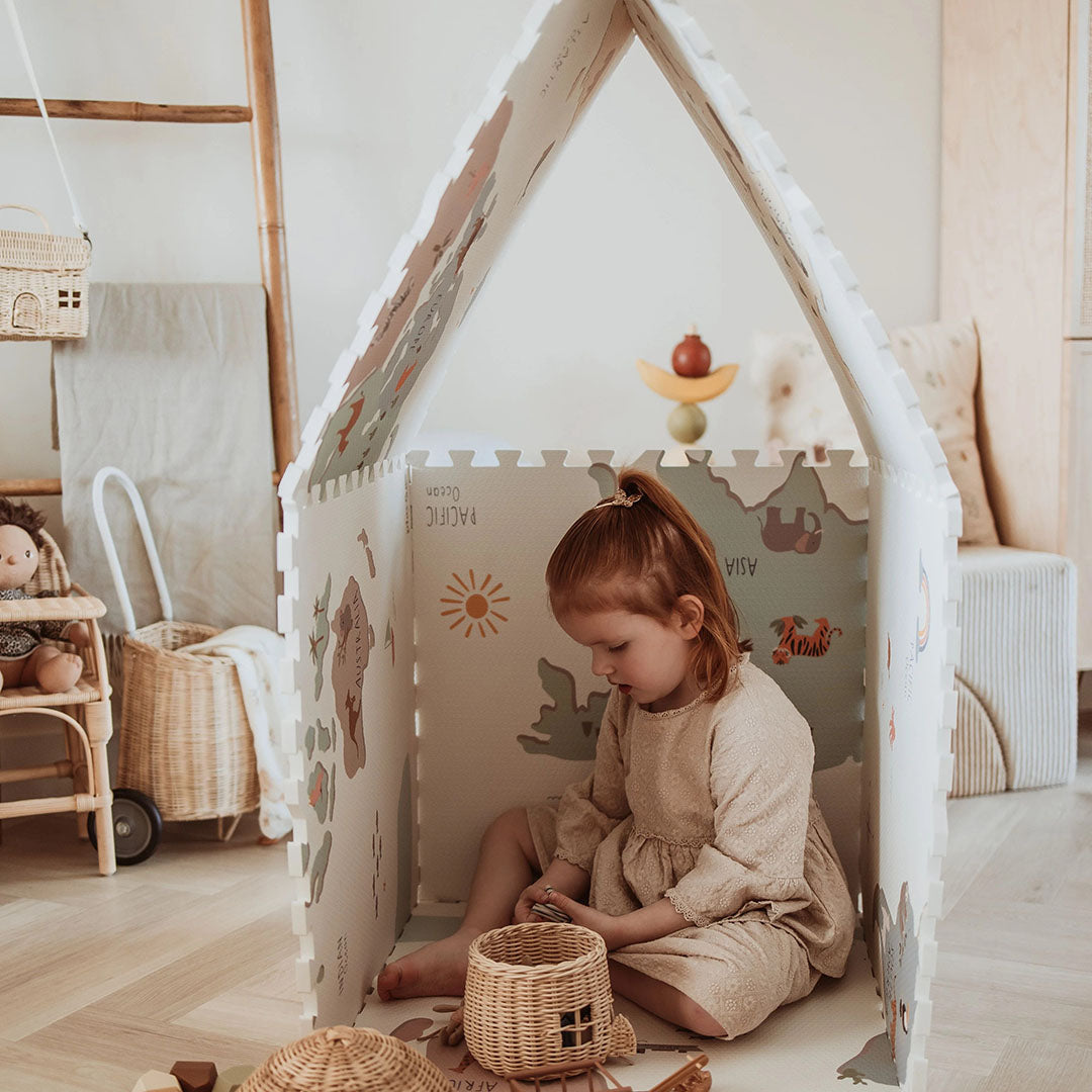 Een kind zit in een huisje gemaakt van play&go foam speelmat tegels met wereldkaartmotief en speelt met een rieten mandje.