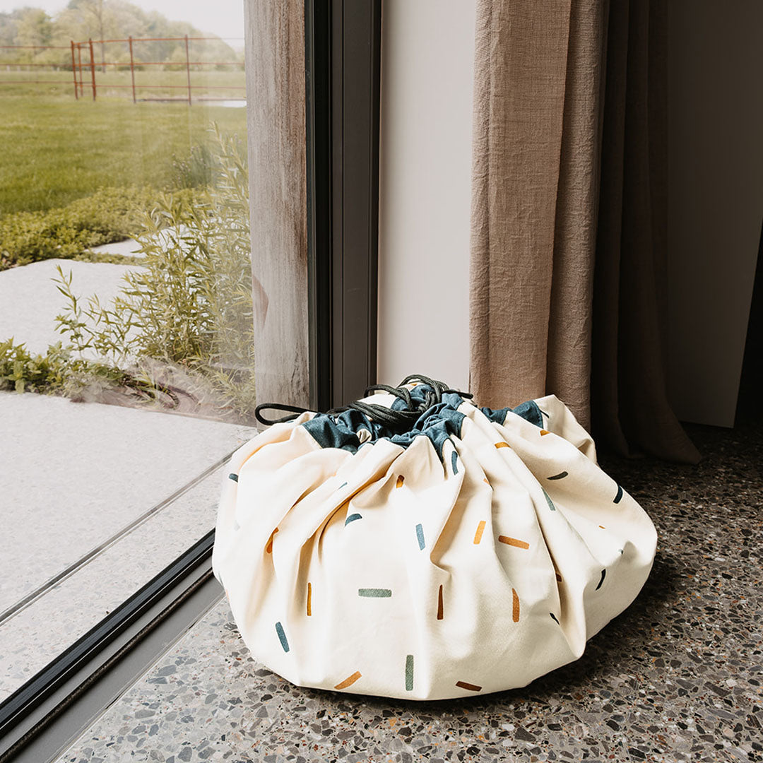 De play&go speelkleed en opbergzak met confetti-patroon staat als een tas op de vloer bij een raam.