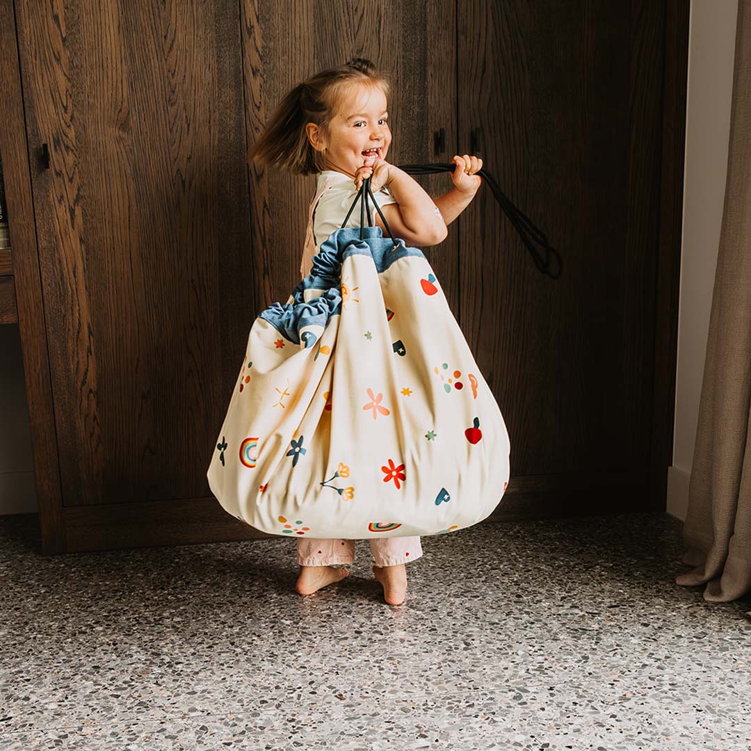 Een lachend kind draagt de play&go speelkleed opbergzak met bloemen, harten en regenbogen als een tas.