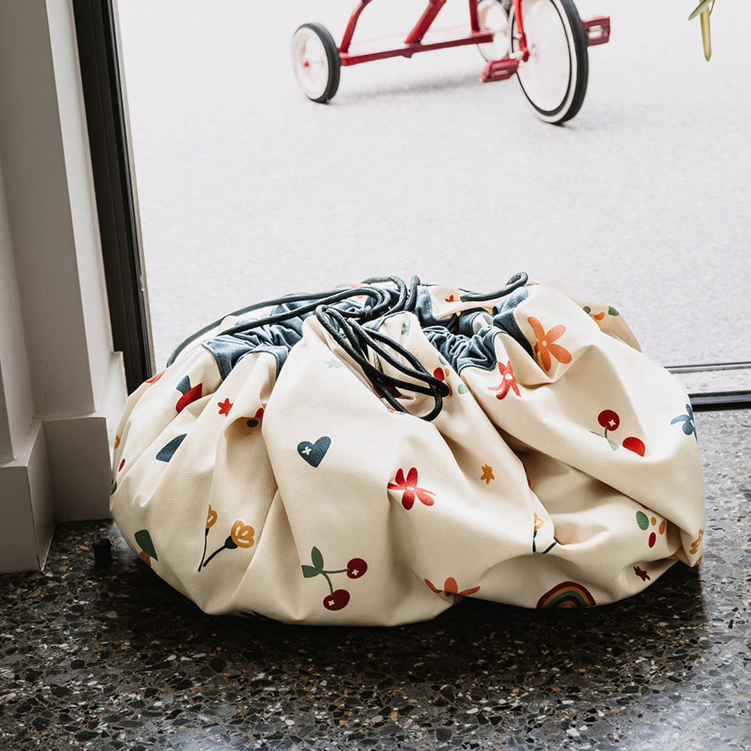 De play&go speelkleed opbergzak met bloemen, harten en regenbogen, dichtgetrokken en vol, staat op de vloer.