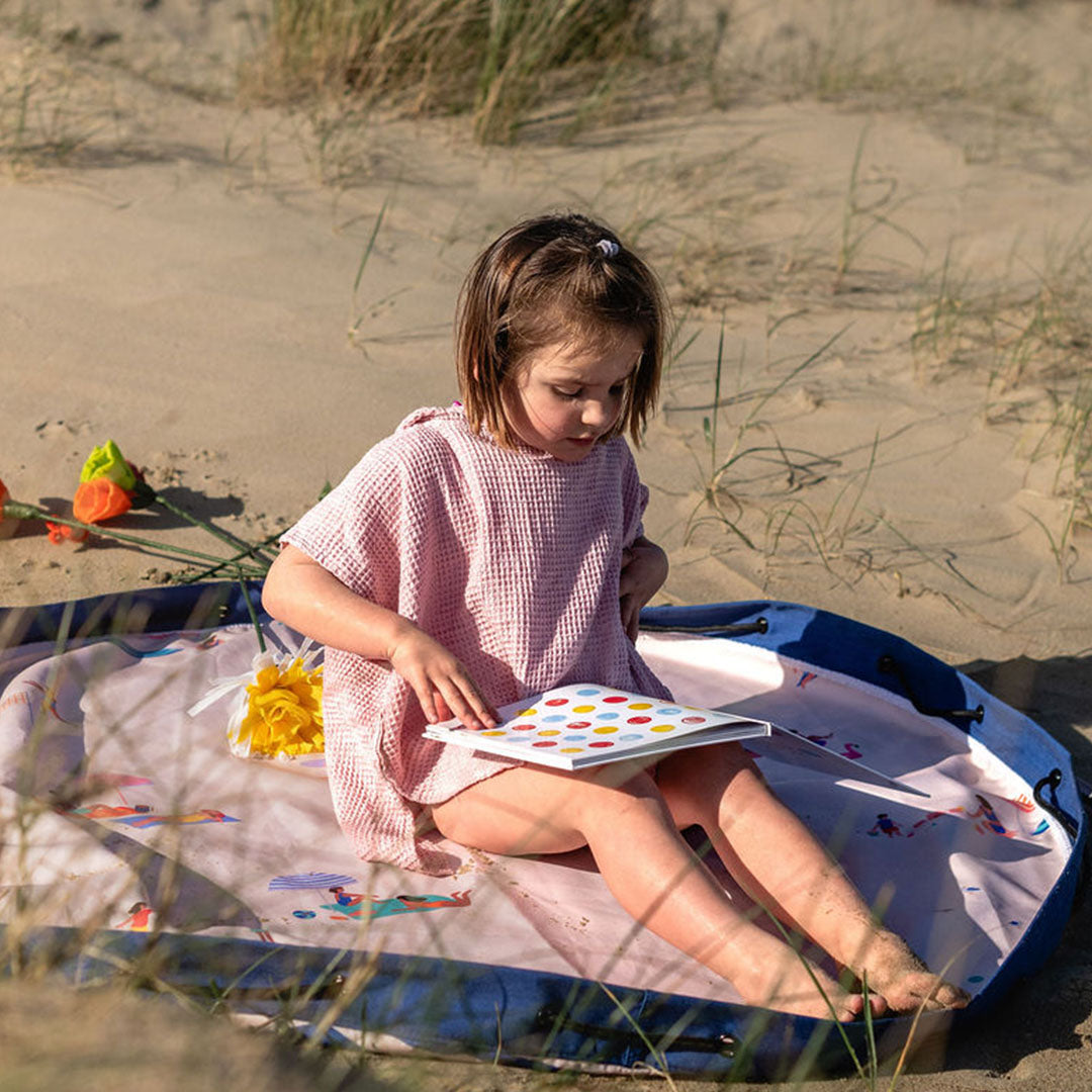 Een kind zit op het play&go speelkleed met strandthema in het zand en leest een boek.