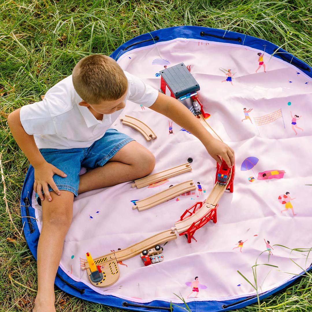 Een jongen speelt met een houten treinbaan op het play&go speelkleed met strandthema in het gras.