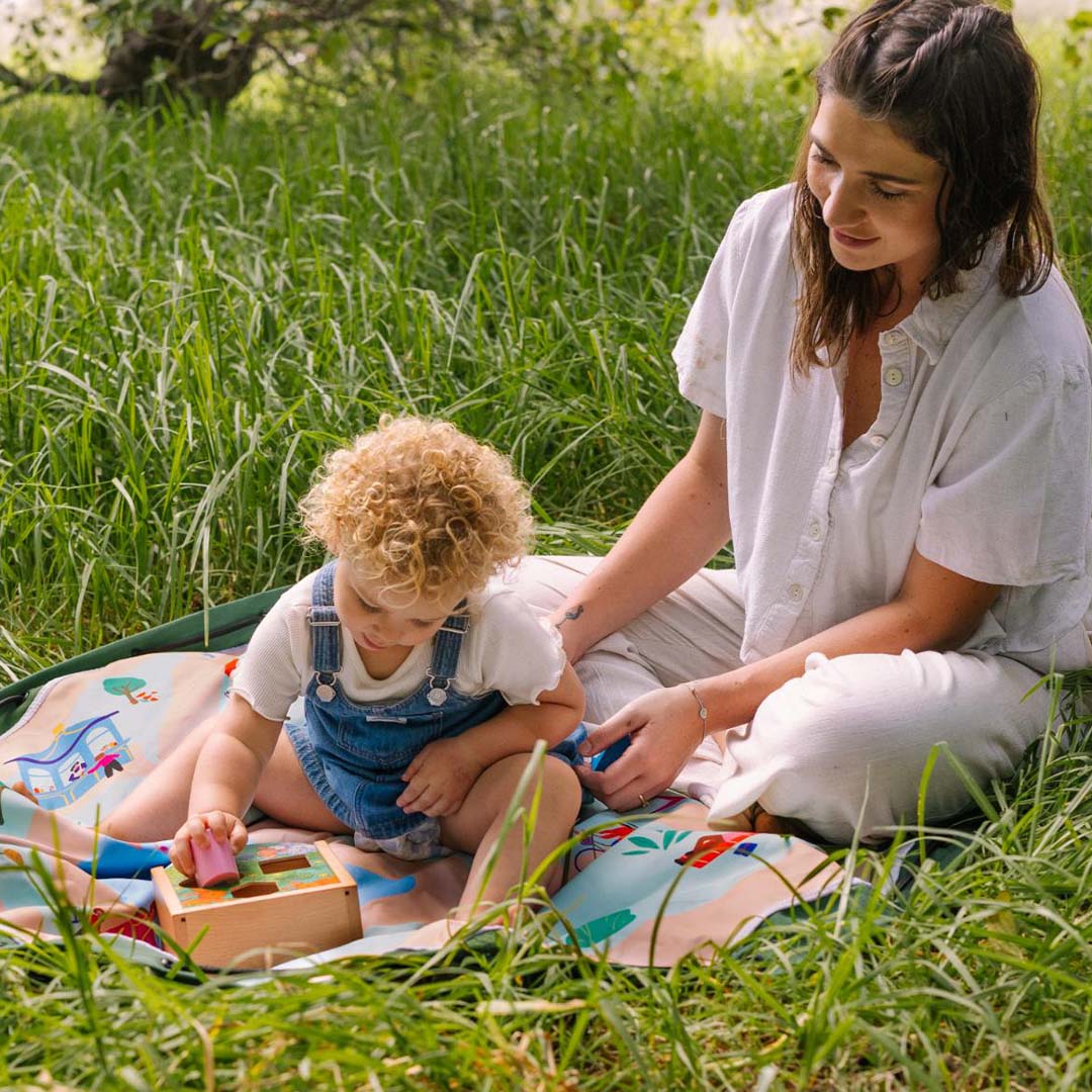 Een vrouw en kind spelen met een houten vormensorteerder op het play&go speelkleed in een grasveld.