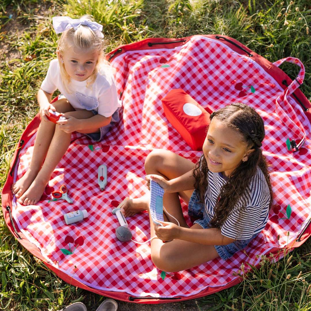 Twee kinderen spelen op een uitgespreid play&go waterdicht speelkleed met rood-wit geruit patroon en kersenmotief in het gras.